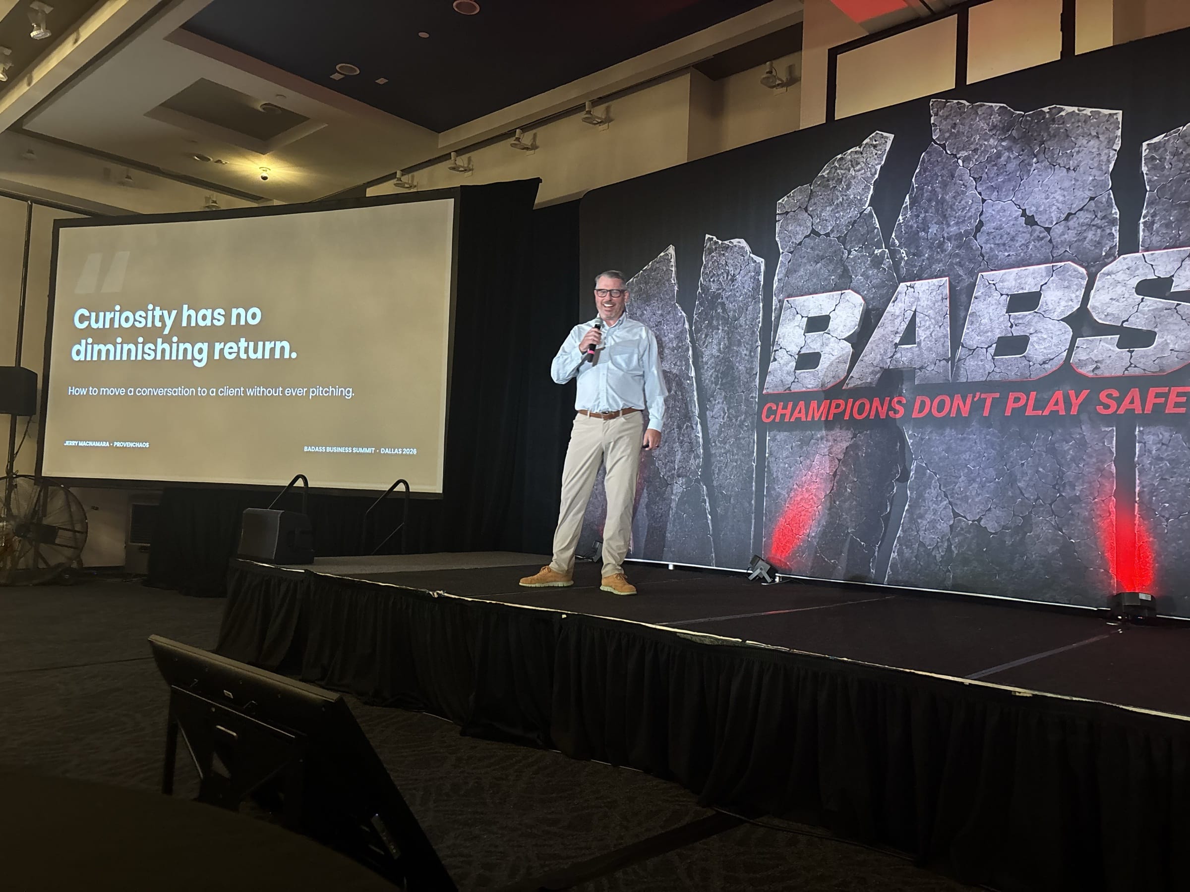 Jerry Macnamara on stage at the Badass Business Summit 2026 in Dallas beside the opening slide of his talk How to Move a Conversation to a Client Without Ever Pitching